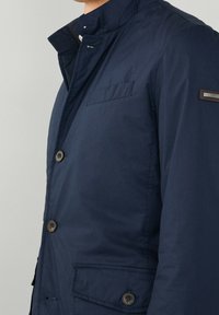 Marinblå jacka med hög krage, knappstängning och bröstficka. Har stora knappar och en märkessymbol på ärmen.