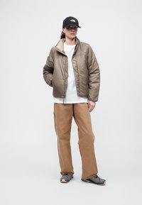 Geacă puf în nuanță deschisă de maro, cu butonare în față, asociată cu pantaloni largi de culoare bej. Cămașă albă dedesubt, încălțăminte gri și o șapcă neagră.