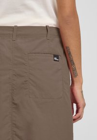 Shorts en tissu marron avec une poche arrière dotée d'une étiquette de logo noire. Texture lisse et design ajusté, portés avec un haut léger.