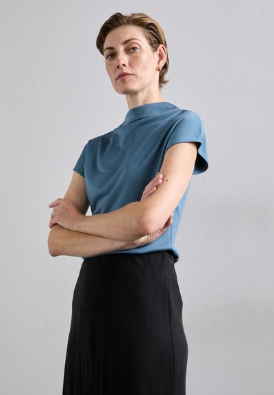 Lyseblå, kortermet topp med høy hals, kombinert med et svart skjørt. Glatt tekstur med minimalistisk design, ingen synlige mønstre eller aksenter.