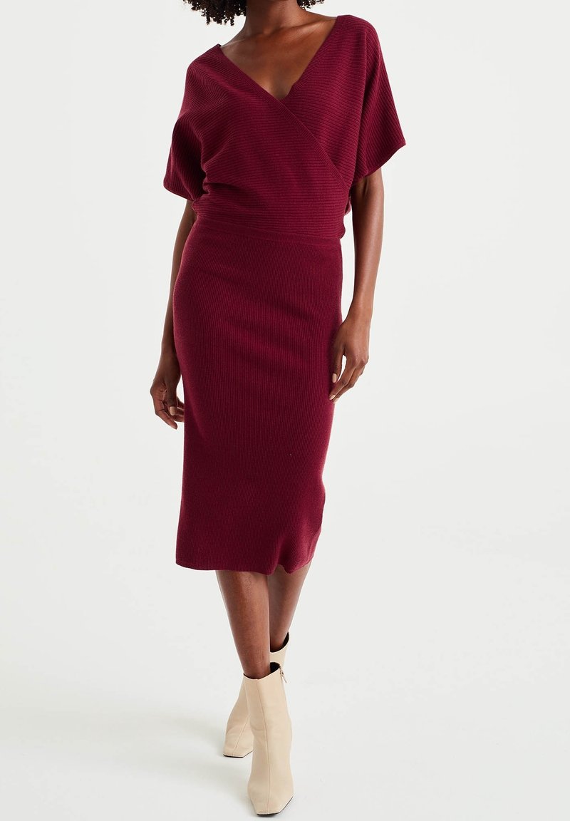 Bordeauxrotes, geripptes Strickkleid mit tiefem V-Ausschnitt, kurzen Ärmeln und figurbetonender Silhouette, das bis zur Mitte der Wade reicht. Kombiniert mit beigen Ankle-Boots.