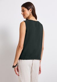 Bezramionkowy, ciemnozielony top z przewiewnego materiału z delikatną teksturą, z dekoltem w kształcie klucza z tyłu, zestawiony z jasnymi spodniami.