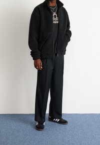 Chaqueta de forro polar negra con cremallera y bolsillos laterales, combinada con pantalones anchos negros y zapatillas negras con rayas blancas.