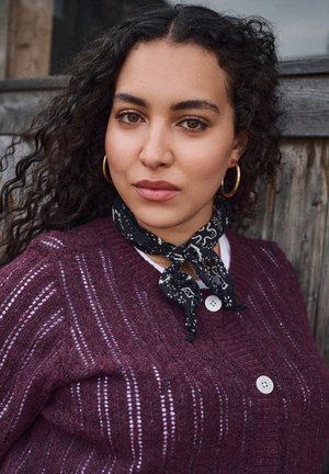 Pull bourgogne avec un tricot texturé, featuring des rayures blanches et des boutons. Écharpe noire à motifs attachée autour du cou, boucles d'oreilles en or.