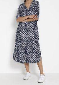 Osoba v námořnických šatech s bílým květinovým potiskem, s polovičními rukávy a bílými teniskami, stojící s překříženýma rukama na bílém pozadí.