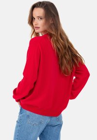 Pull rouge surdimensionné avec un col arrondi et des manches longues. Texture douce, poignets et ourlet côtelés. Porté avec un jean bleu clair.