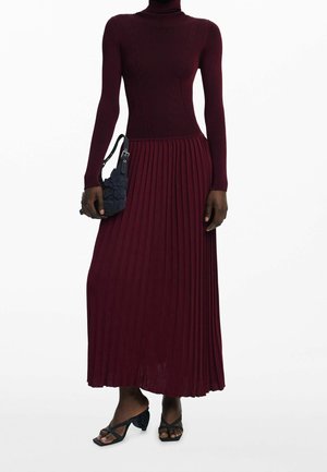 Mujer con un vestido plisado burdeos de manga larga, sosteniendo un bolso negro con textura y llevando sandalias negras de tacón.
