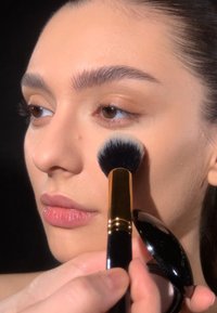 Make-up-Anwendung mit einem großen, weichen Pinsel mit schwarzem Griff und goldenen Akzenten, der eine glatte, helle Foundation auf der Haut hervorhebt.