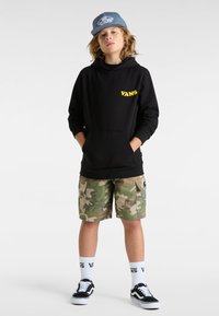 Felpa nera con cappuccio e logo giallo "VANS", pantaloni cargo mimetici, sneakers nere, calzini bianchi con testo "VANS", cappellino blu.