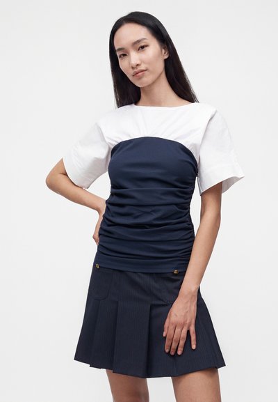 Femme aux longs cheveux noirs portant un haut blanc à manches courtes sous un bustier bleu marine froncé et une jupe plissée bleu marine avec des boutons dorés.