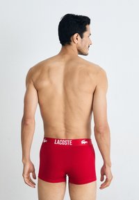 Red cotton boxer briefs with a white "LACOSTE" logo on the waistband. Form-fitting design, smooth texture, and mid-thigh length.