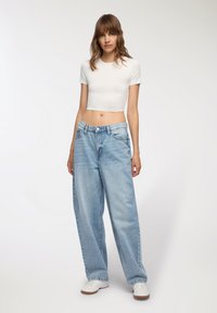 T-shirt bianca cropped, jeans in denim oversize blu chiaro con chiusura a bottoni, e sneakers bianche con suola texturizzata.