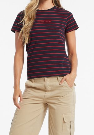 Femme portant un t-shirt Guess Jeans rayé bleu marine et rouge, associé à un pantalon cargo beige, les mains reposant dans et près des poches.