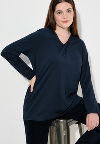 Marinblå långärmad topp med V-ringning, featuring ett draperat framstycke och mjuk textur. Matchad med mörka byxor och sitter på en silverfärgad metallpall.