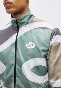 Άνδρας με ζακέτα με φερμουάρ σε πράσινο, άσπρο, γκρι και μπεζ με αφηρημένα μοτίβα και λογότυπο EA7 Emporio Armani στο στήθος.