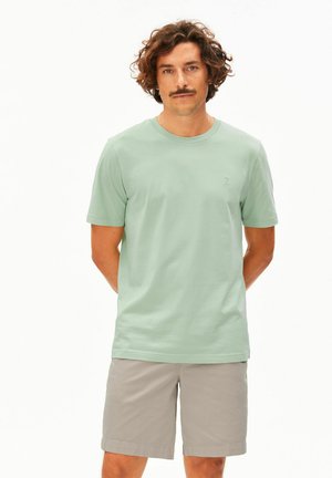 Mann mit lockigem braunem Haar und Schnurrbart, der ein hellgrünes kurzärmeliges T-Shirt und beige Shorts trägt und mit den Händen hinter dem Rücken steht.