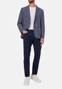 Blazer bleu marine en tissu texturé, design à un seul bouton, avec deux boutons. Associé à un t-shirt blanc, un pantalon sombre et des baskets blanches.