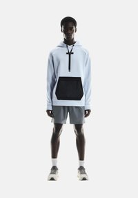 Light blue hooded sweatshirt with black pocket; features drawstring and textured fabric. Paired with gray shorts and chunky sneakers.