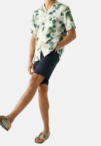 Koszulka z krótkim rękawem na jasnym kremowym tle, zdobiona zielonymi wzorami palm i kwiatów. Połączona z granatowymi szortami i beżowymi sandałami.