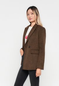 Rjava tweed blazer z odrezanimi reverji, enim gumbom in dvema sprednjima žepoma. V kombinaciji z belo srajco in črnimi hlačami.