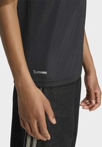 Asmuo dėvi juodą Climacool marškinius ir tamsias kelnes, rankos atsipalaidavusios šonuose, stovintis priešais vientisą foną.