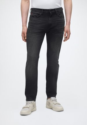 Άνδρας που φορά στενά μαύρα τζιν και λευκά αθλητικά παπούτσια, στέκεται μπροστά σε απλό λευκό φόντο.