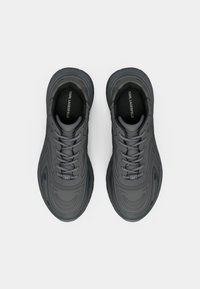 Zapatos deportivos grises con cordones, parte superior texturizada, líneas de diseño fluidas y suelas de goma con patrón. Presentan el logo en el talón y la lengüeta.