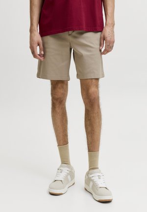 Person trägt beigefarbene Shorts, ein dunkelrotes Shirt, beigefarbene Socken und weiße Sneaker und steht vor einem schlichten, hellen Hintergrund.