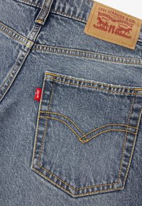 Μπλε τζιν παντελόνια με κίτρινη ραφή, χαρακτηριστικό σχέδιο πίσω τσέπης και καφέ δερμάτινη ετικέτα με το λογότυπο της Levi Strauss.