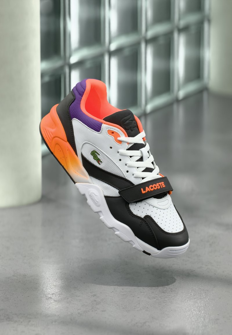 Baskets Lacoste présentant un design blanc, noir et orange avec un talon violet. Comprend des détails perforés et une sangle Velcro.