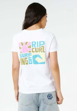 Femme aux longs cheveux foncés vue de dos, portant un t-shirt blanc avec un motif coloré "Rip Curl Surfing Co" et un jean bleu clair avec un patch logo.