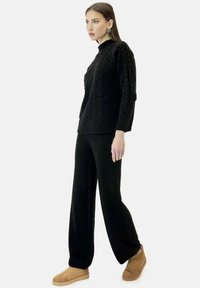 Suéter negro de punto con patrón de trenza texturizada, combinado con pantalones anchos negros y zapatos de lona marrones. Modelo de pie frente a un fondo liso.