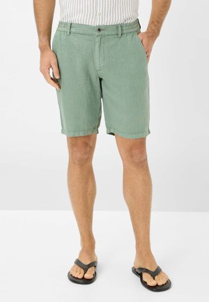 Shorts in lino verde con vita elastica, chiusura con bottone e tasche laterali. Indossati con infradito nere su uno sfondo neutro.
