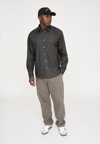 Schwarz gestreifte Hemd mit Knöpfen, graue Hose und weiße Sneakers mit schwarzen Akzenten. Die Person trägt eine schwarze Kappe mit goldfarbenem Text.