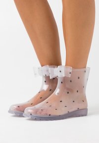 Clear, polka-dotted rain boots with a translucent design, featuring a bow accent at the ankle and a textured rubber sole.