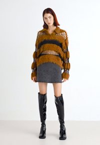 Pulover fuzos cu dungi maro și negre, fustă mini neagră cu un model de puncte și cizme negre lucioase până la genunchi pe un model, pe un fundal alb.