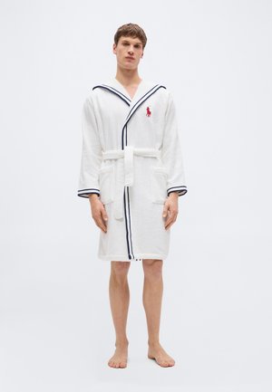 Uomo in piedi a piedi nudi che indossa un accappatoio bianco con bordi blu navy e un logo rosso sul petto, mani rilassate ai lati.