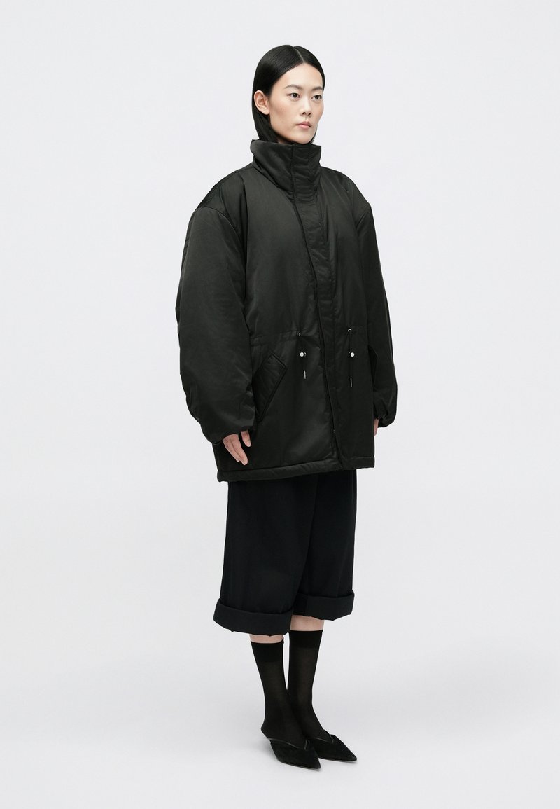 Black puffer jacket with high collar, large sleeves, and two front pockets. Paired with dark, wide-leg trousers and pointed black shoes.