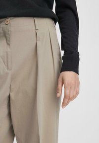 Beige plooibroek met een relaxte pasvorm, voorzien van tan knopen en een gladde, licht gestructureerde stof. Zwarte trui-mouw zichtbaar.