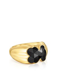 Anello d'oro con una fascia larga, caratterizzato da una pietra preziosa nera a forma di farfalla, sfaccettata e incastonata sopra, che riflette la luce su una superficie bianca.