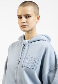 Sweat à capuche bleu clair en tissu doux, avec un design brodé décoloré '100' sur la poitrine gauche. Fermeture éclair à l'avant, coupe décontractée.