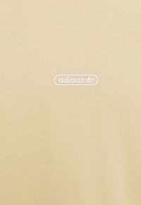Ljusgul bomullströja med en vit broderad Adidas-logotyp i en rundad rektangulär form nära nedre högra hörnet.
