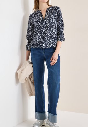 Woman wearing a navy patterned blouse, cuffed blue jeans, silver sneakers, holding a beige jacket against a light background.