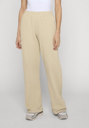 Persona che indossa pantaloni beige a gamba larga, top bianco e scarpe da ginnastica grigie, in piedi contro uno sfondo chiaro e uniforme.