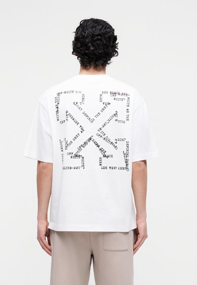 Άνδρας που φοράει λευκό φαρδύ T-shirt με αφηρημένο μαύρο κείμενο που σχηματίζει σχήματα στην πλάτη, συνδυασμένο με μπεζ παντελόνι σε απλό φόντο.