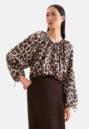 Frau trägt eine lockere Bluse mit Leopardenmuster und langen Puffärmeln sowie einen dunkelbraunen, hoch taillierten Rock und steht mit einer Hand in der Hüfte.