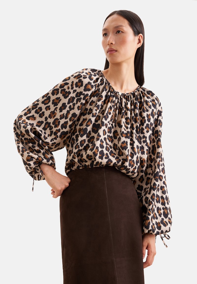 Femme portant un chemisier ample à imprimé léopard avec de longues manches bouffantes et une jupe taille haute marron foncé, debout avec une main sur la hanche.