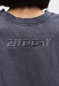 Κοντινό πλάνο της πλάτης ενός ατόμου με σκούρο γκρι πουκάμισο που έχει ανάγλυφο μοβ κείμενο "2ND DAY" κοντά στη γραμμή του λαιμού.