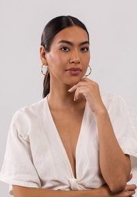 Goldene Creolen mit strukturiertem Finish, kombiniert mit einem weißen Leinenoberteil mit geknoteter Front. Ein glatter Hautton verstärkt das Gesamtbild.