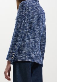 Femme portant un blazer à carreaux bleu et blanc texturé avec des boutons argentés sur la manche, associé à un jean en denim bleu foncé.